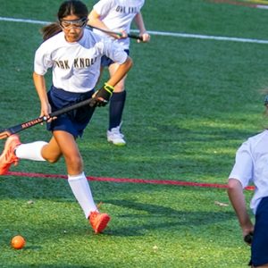 Lower School Field Hockey athlete plays the ball in front of a teammate and an opposing player.