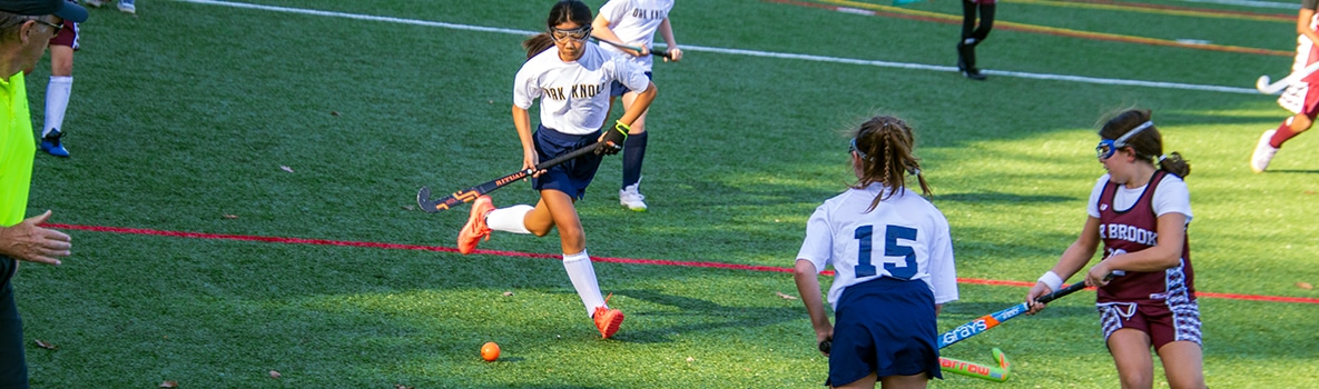Lower School Field Hockey athlete plays the ball in front of a teammate and an opposing player.